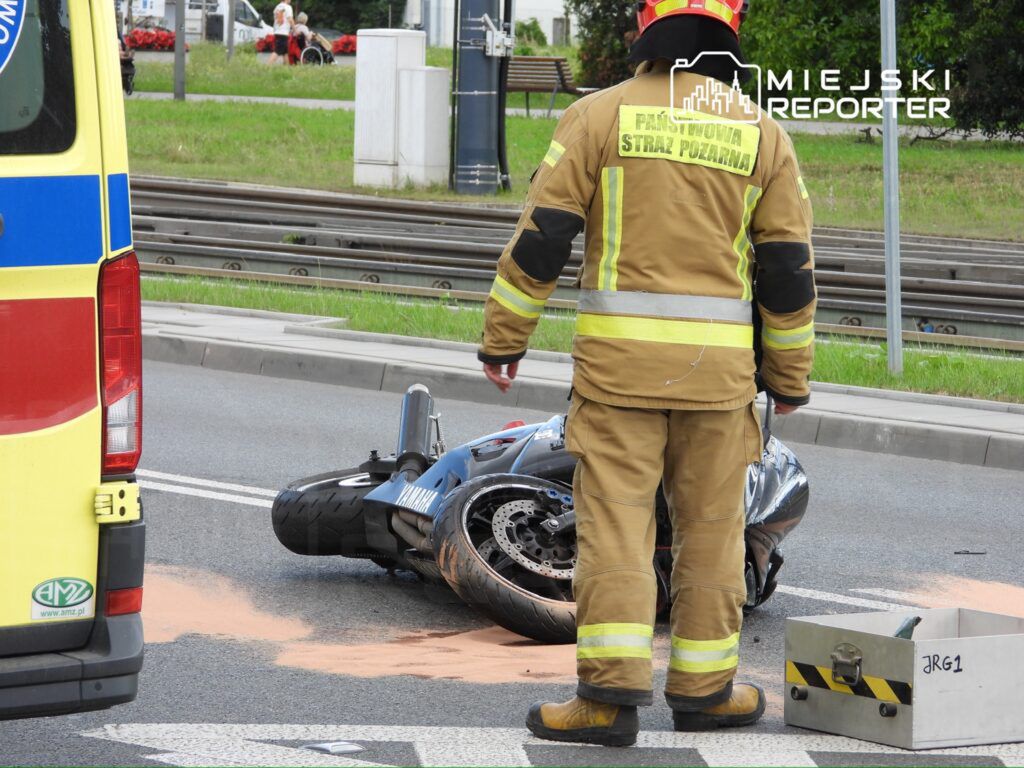 Strażak w mundurze z logo Państwowej Straży Pożarnej stoi obok przewróconego motocykla na drodze.