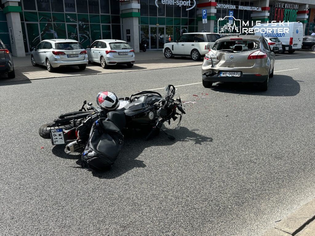 Motocykl leży na jezdni obok uszkodzonego samochodu osobowego, w tle widać parking z innymi pojazdami.