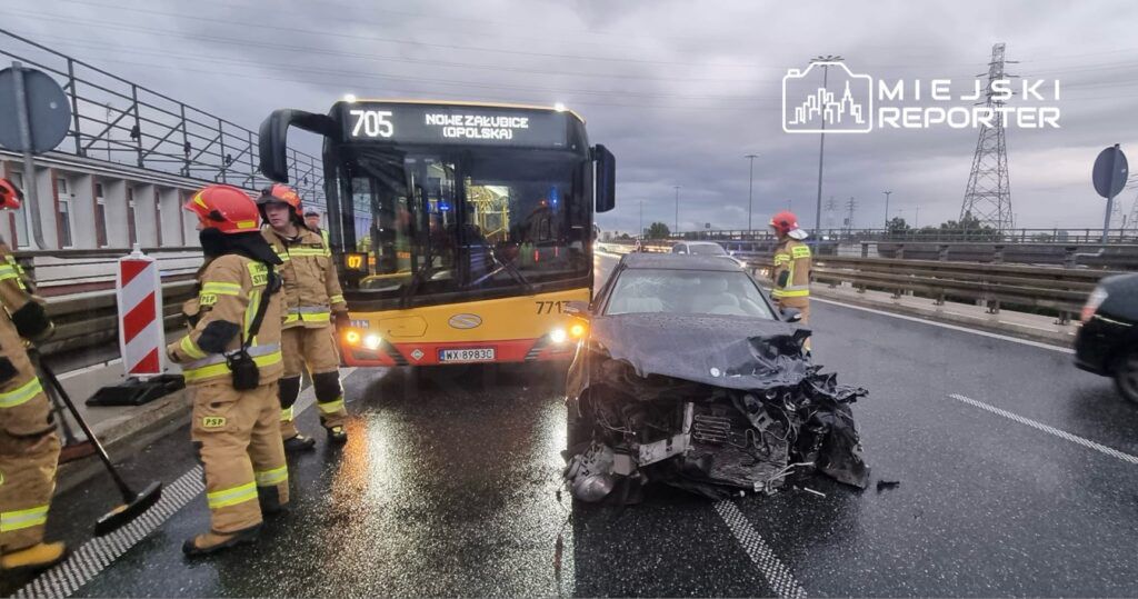 Grupa strażaków w mundurach zabezpiecza miejsce wypadku drogowego z uszkodzonym samochodem osobowym obok autobusu.