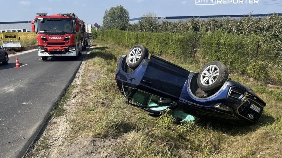 Na poboczu drogi leży przewrócone na dach czarne auto, obok stoi wóz strażacki. W tle widoczne są krzewy.