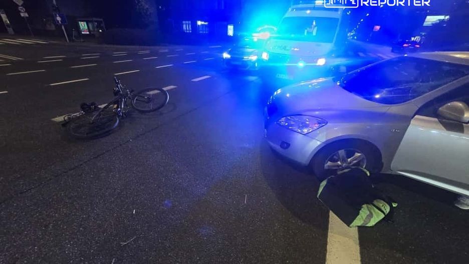 Na jezdni leży przewrócony rower obok srebrnego samochodu, a w tle widać pojazd ratunkowy z włączonymi sygnałami świetlnymi.