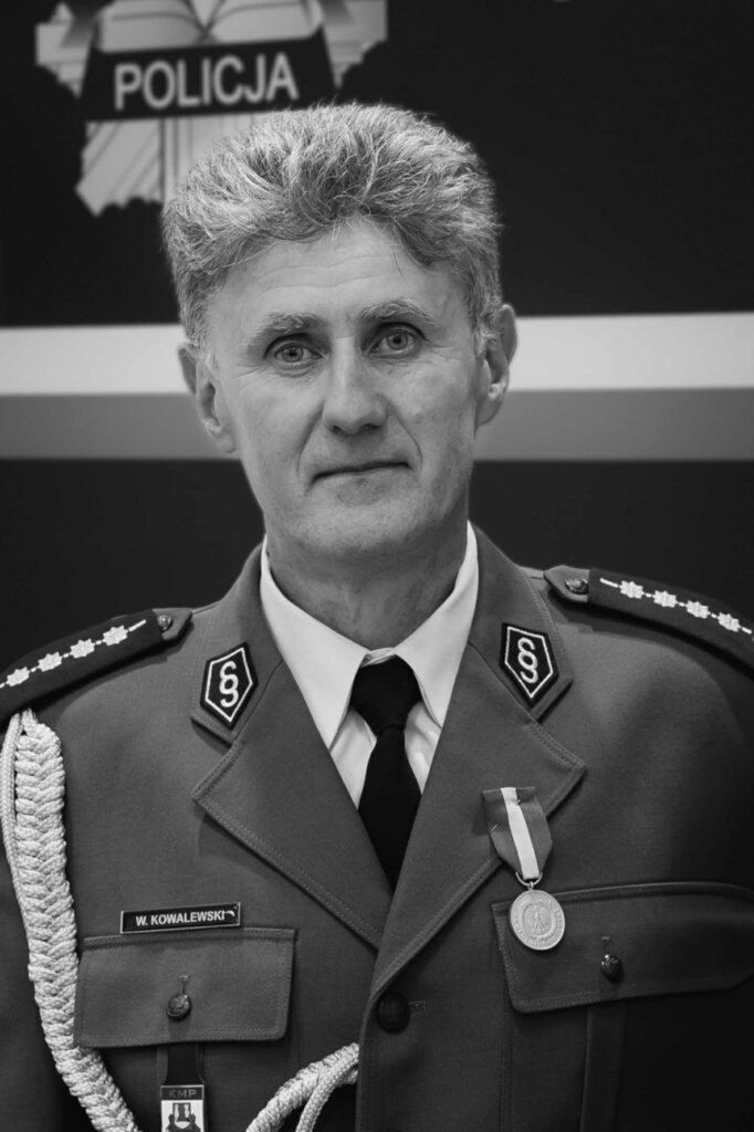 Mężczyzna w mundurze policyjnym z medalem na piersi, pozujący przed tłem z napisem "POLICJA".