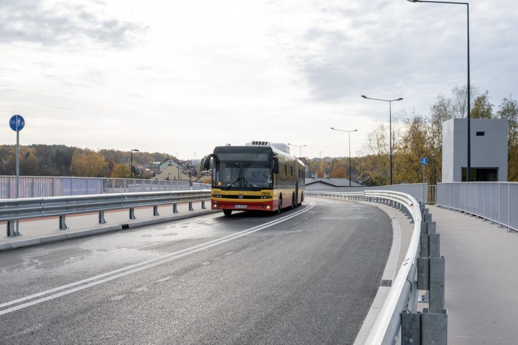 Autobus zjeżdża z nowego mostu na ulicę, mijając oznaczenie dla rowerzystów i otoczenie z drzewami w tle.