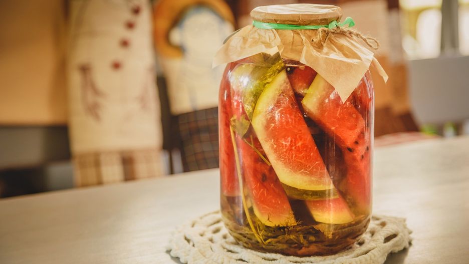 Marinated or pickled watermelon with herbs in a glass jar on a wooden table in a kitchen.