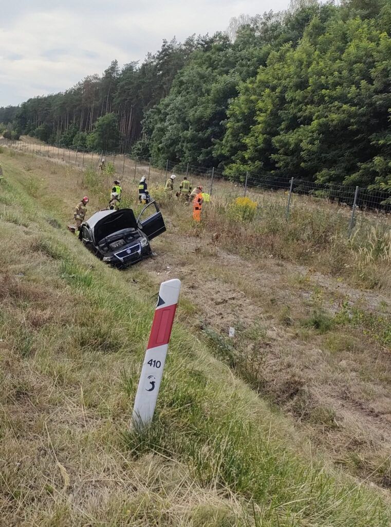 Grupa ratowników w odzieży ochronnej przeszukuje teren przy przewróconym samochodzie na poboczu drogi, otoczonym drzewami.