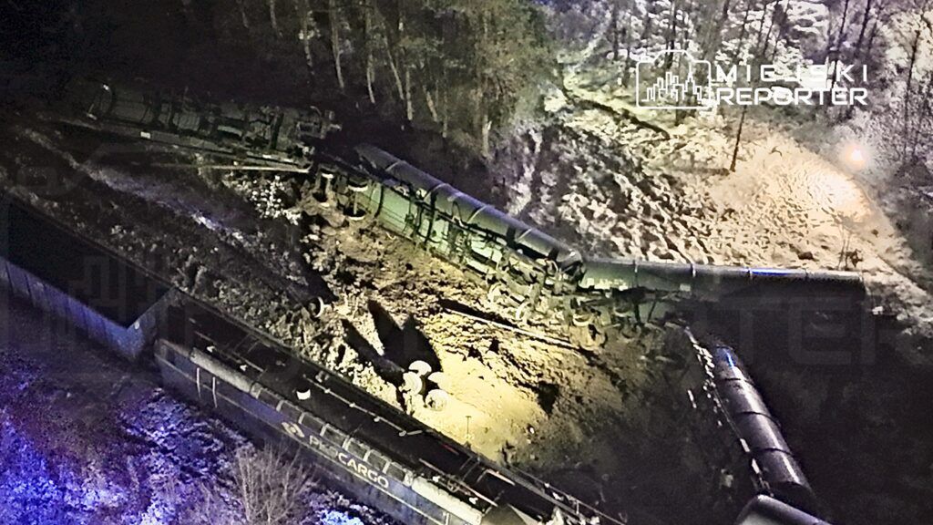 Złomowany pociąg towarowy leży na boku w zalesionym terenie, otoczony śniegiem i ciemnością.