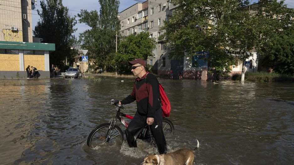Lokalny mieszkaniec z rowerem i psem idzie ulicą obok budynków w Chersoniu na Ukrainie, wtorek, 6 czerwca 2023 r., które zostały zalane po wysadzeniu tamy Kachowka w nocy.
