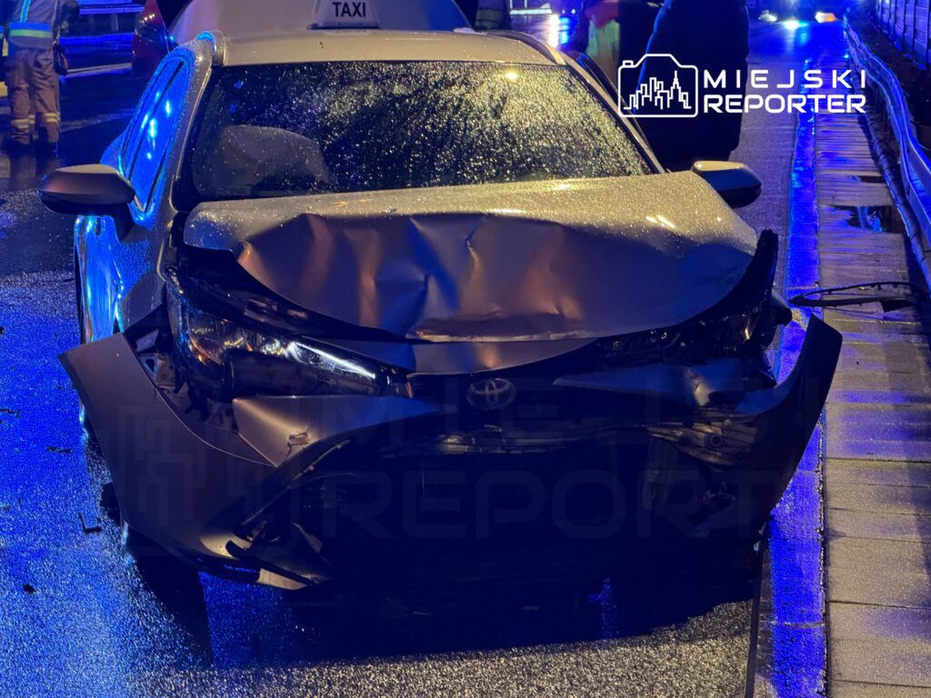 Uszkodzony samochód osobowy z wgniecionym przodem stoi na mokrej nawierzchni, otoczony przez służby ratunkowe.