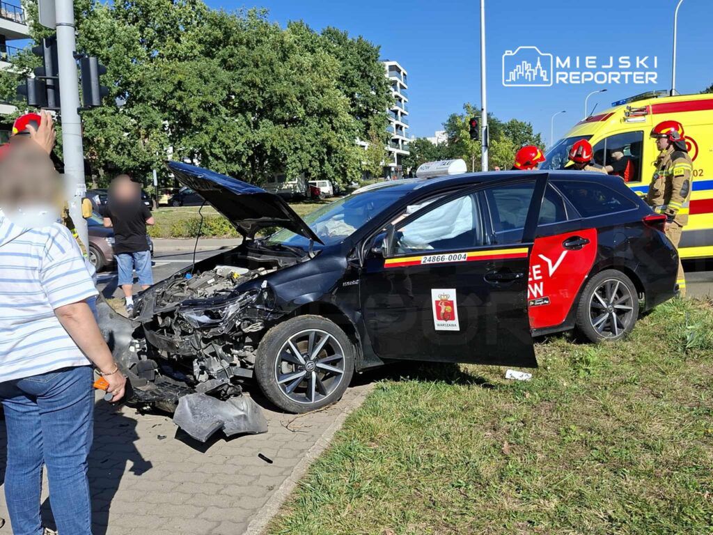 Czarny samochód osobowy z uszkodzoną przednią częścią stoi na poboczu, obok grupy osób oraz wozu strażackiego.