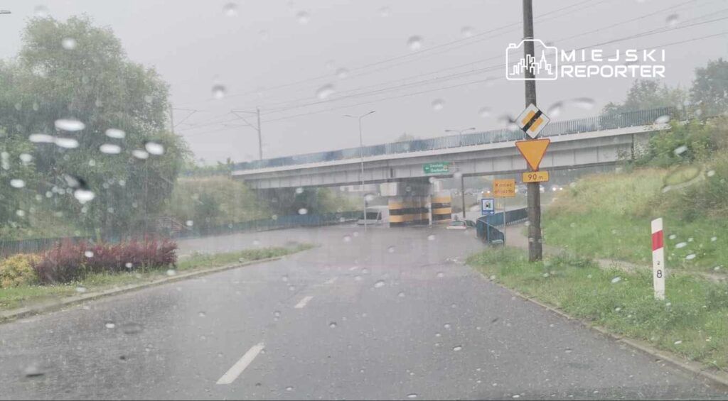 Intensywna ulewa powodująca podtopienie drogi w okolicy wiaduktu, widoczne kałuże i zamglony krajobraz.