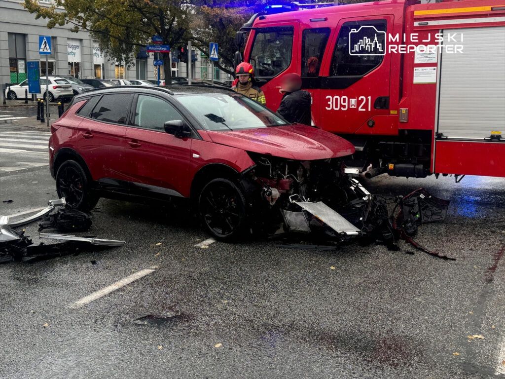 Czerwony samochód osobowy z uszkodzoną przednią częścią stoi na mokrej ulicy obok wozu strażackiego.
