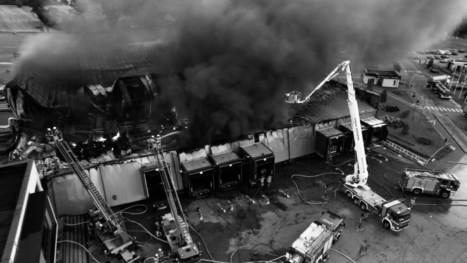 Wysoka chmura czarnego dymu unosząca się z płonącego budynku, w tle strażacy używający podnośnika do gaszenia ognia.