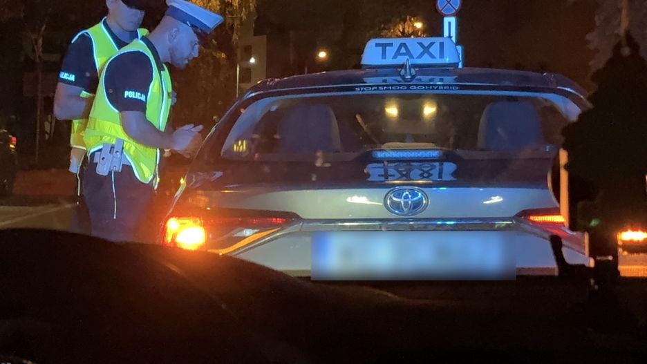 Dwaj policjanci w kamizelkach odblaskowych kontrolują taksówkę na ulicy w nocy.