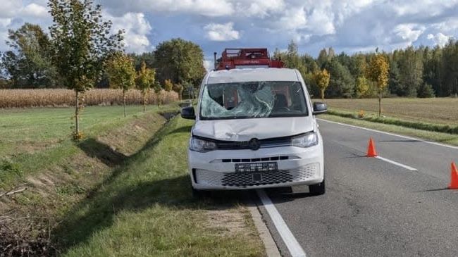 Biały samochód dostawczy z uszkodzoną szybą stoi na poboczu drogi, obok pomarańczowe trójkąty ostrzegawcze.
