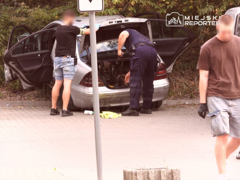 Mężczyzna w mundurze przeszukuje bagażnik samochodu, podczas gdy inny mężczyzna obserwuje sytuację w tle.