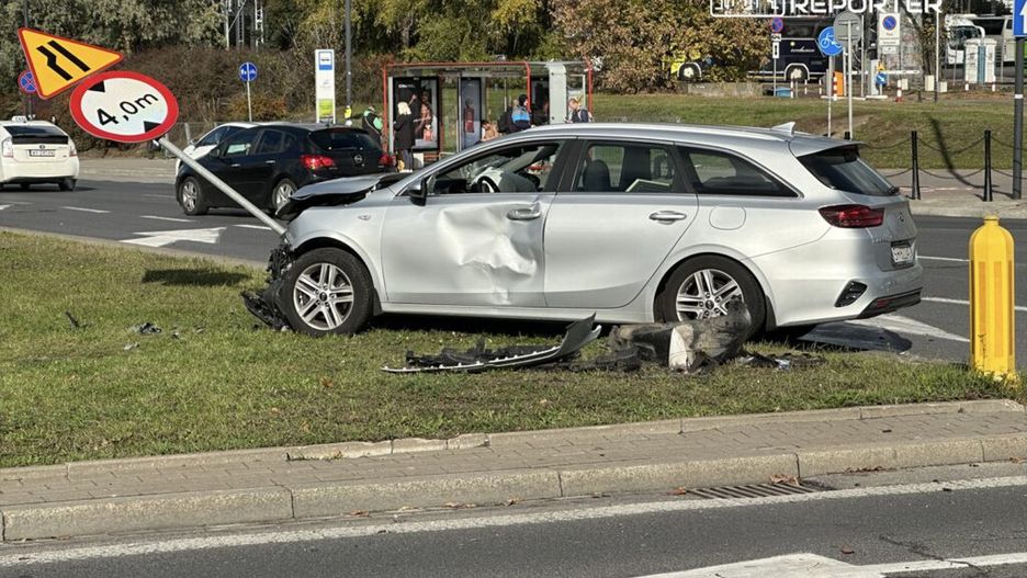Uszkodzony srebrny samochód osobowy zaparkowany na trawie obok znaku drogowym informującym o ograniczeniu wysokości do 4 m.