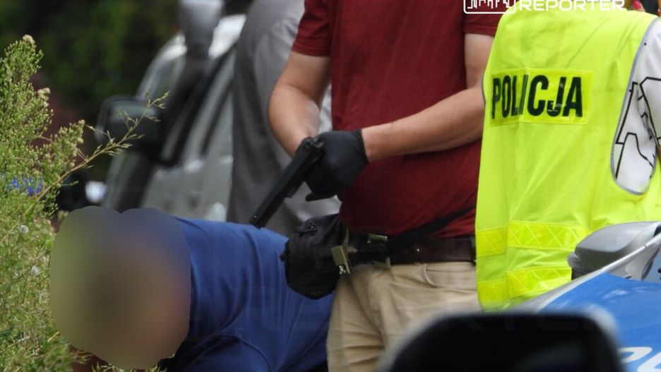 Mężczyzna w czerwonej koszulce trzyma broń, podczas gdy inny mężczyzna klęczy obok policyjnego samochodu.