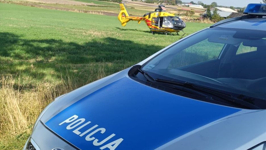 Policyjny samochód z widocznym napisem "POLICJA" stoi na zielonej łące obok lądowiska dla helikopterów.