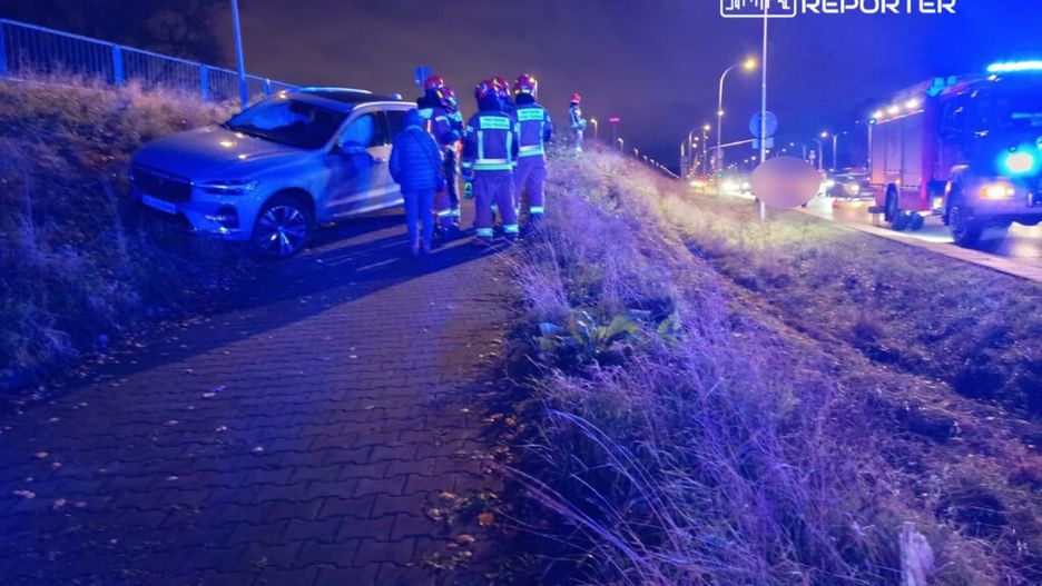 Grupa strażaków rozmawia z osobą obok uszkodzonego samochodu na poboczu drogi w nocy.