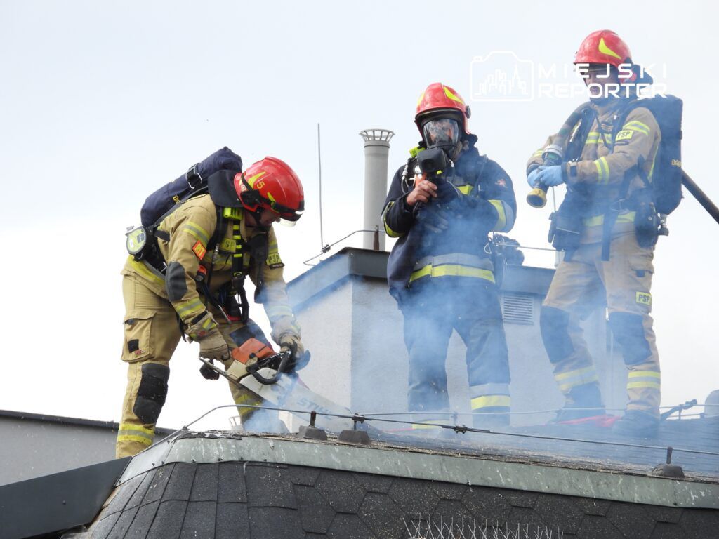 Strażacy w hełmach i ubraniach ochronnych usuwają dachówki z dachu, w tle unosi się dym.