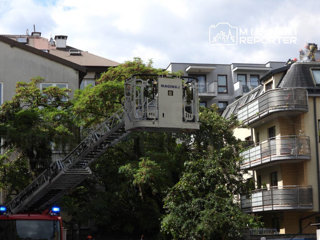 Wóz strażacki z drabiną podjeżdża pod budynek mieszkalny, a strażacy pracują na dachu.