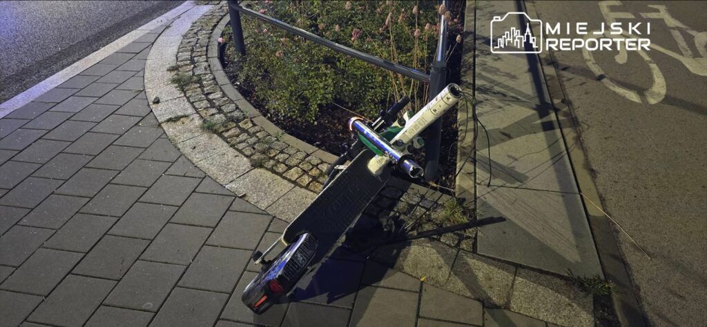 Złamana hulajnoga elektryczna oparta o metalowy płot w oświetlonym nocą otoczeniu. W tle widoczne rośliny i chodnik.