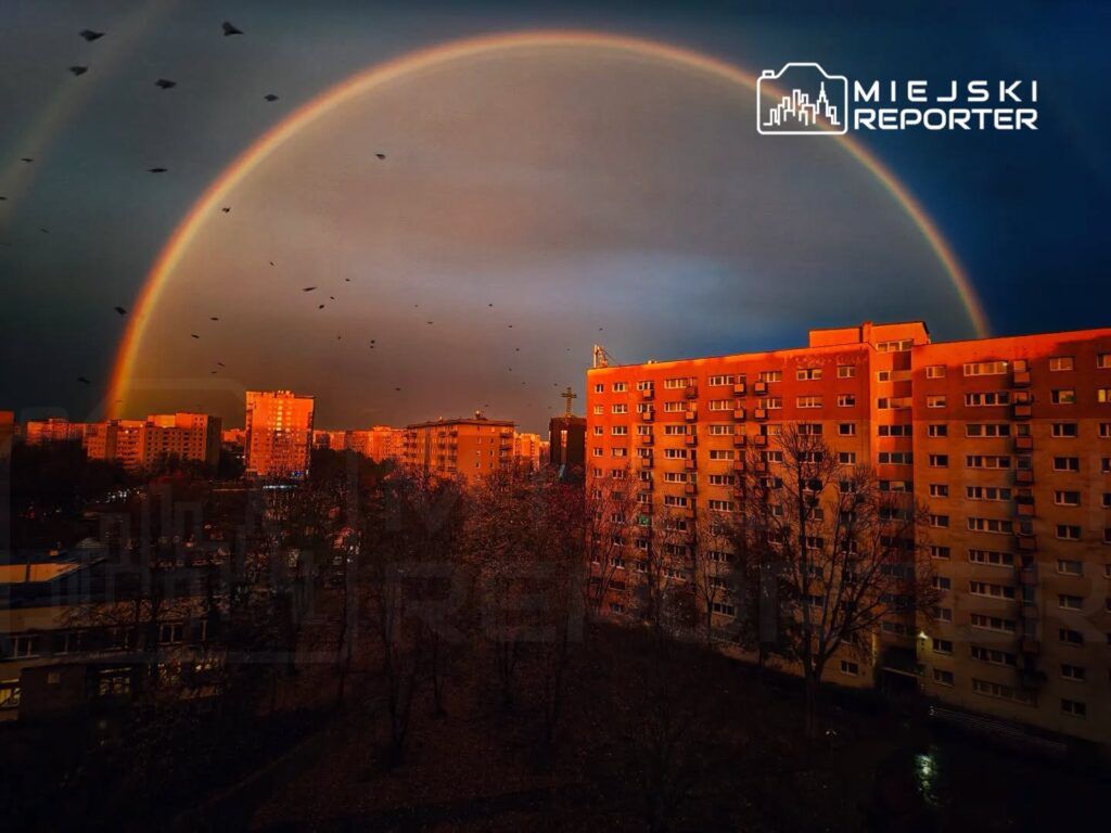 Tęcza rozciągająca się nad osiedlem mieszkaniowym podczas zachodu słońca, w tle ciemne chmury i ptaki w locie.