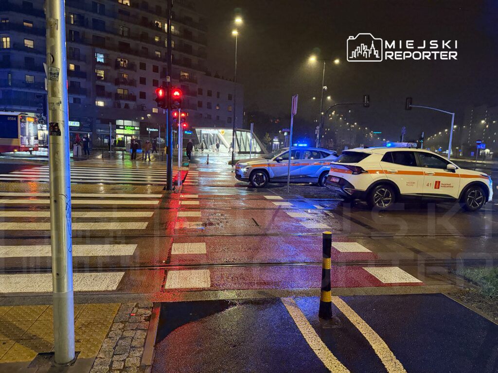 Policyjny radiowóz z włączonymi sygnałami świetlnymi zatrzymany na skrzyżowaniu w deszczowy wieczór.