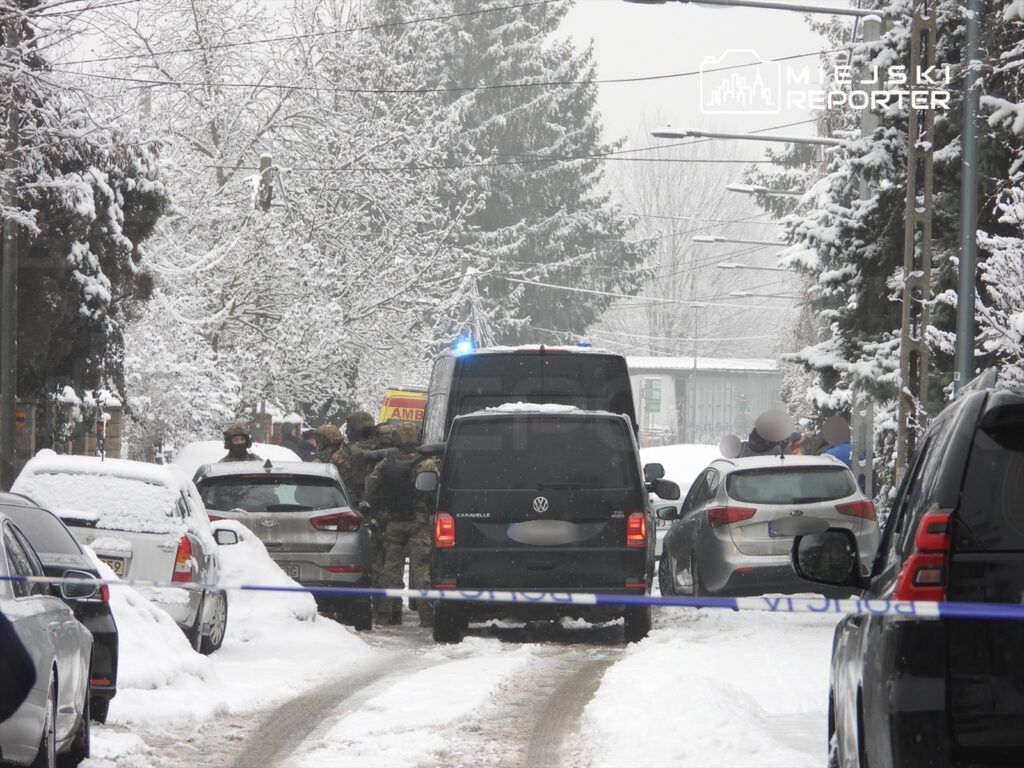 Grupa funkcjonariuszy w kamizelkach taktycznych przeszukuje teren w zaśnieżonym otoczeniu ulicy, obok zaparkowanych samochodów.