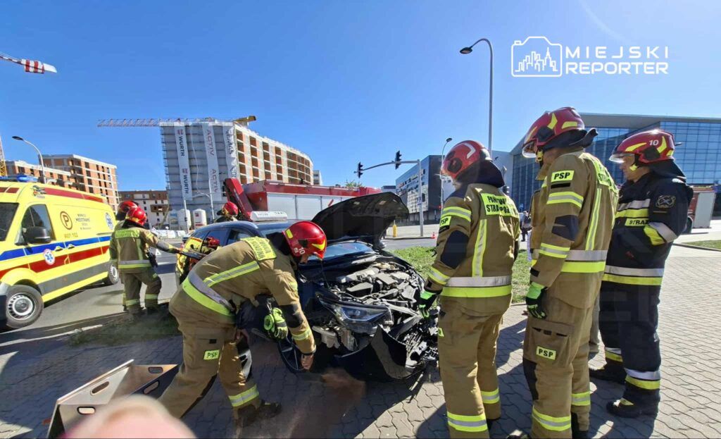 Grupa strażaków w mundurach ratunkowych pracuje przy uszkodzonym samochodzie na ruchliwej ulicy.
