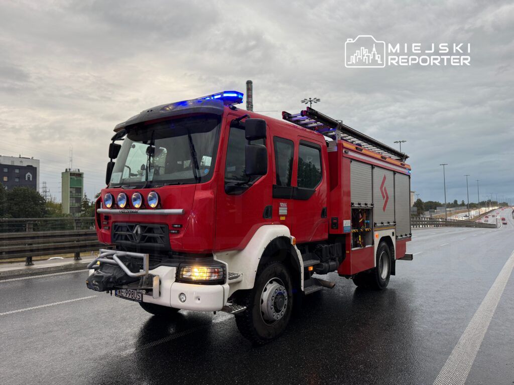 Czerwony wóz strażacki z włączonymi sygnałami świetlnymi stoi na mokrej jezdni w miejskim otoczeniu.