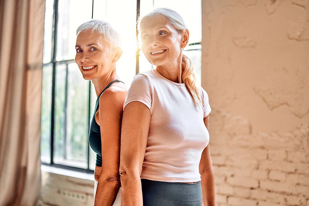 Dwie dojrzałe kobiety stoją plecami do siebie i uśmiechają się w jasnym wnętrzu przy dużym oknie. - zdjęcie