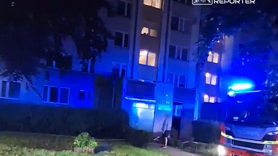 Straż pożarna z włączonymi sygnałami świetlnymi stoi przed budynkiem mieszkalnym w nocy, obok widoczna osoba.
