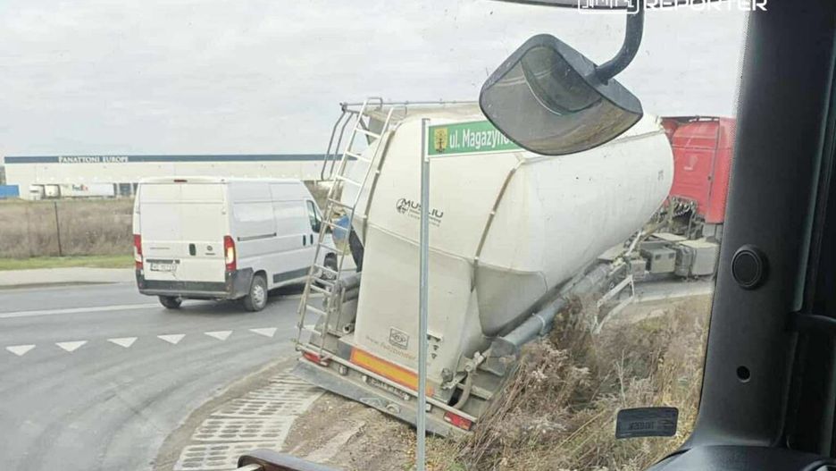 Ciężarówka z cysterną utknęła na poboczu drogi, obok białego furgonu w okolicy przemysłowej.