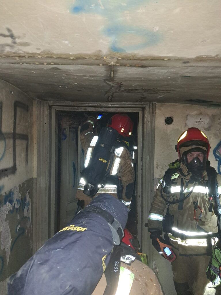 Grupa strażaków w pełnym wyposażeniu przeszukuje zniszczony korytarz w opuszczonym budynku.