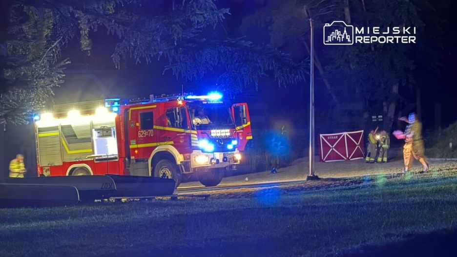 Czerwony wóz strażacki z włączonymi sygnałami świetlnymi stoi na drodze, obok dwóch strażaków w mundurach.