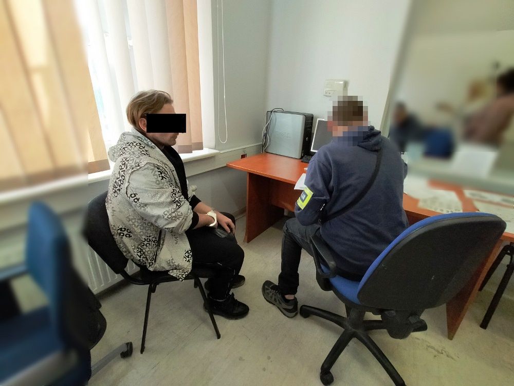 Mężczyzna w jasnej kurtce siedzi na krześle, rozmawiając z osobą w niebieskiej bluzie siedzącą przy biurku w biurze.