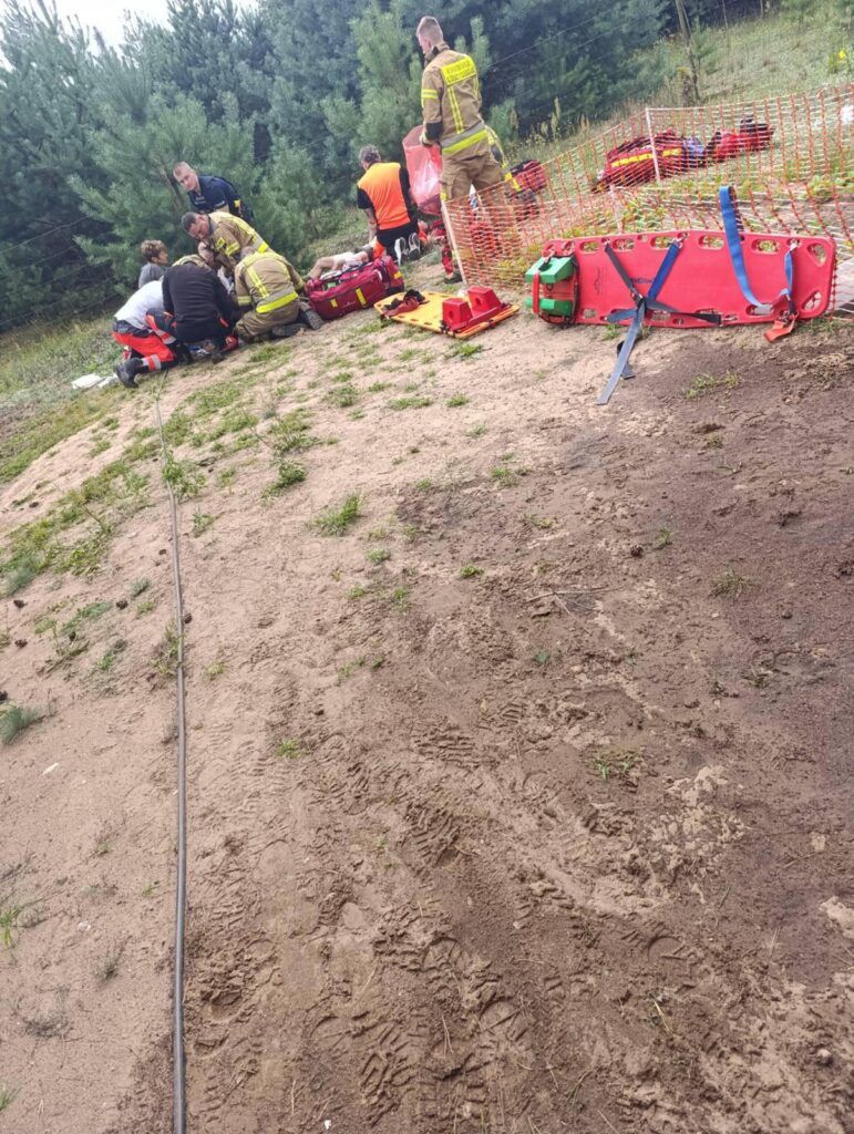 Grupa ratowników i strażaków udziela pomocy osobie leżącej na ziemi w lesie, otoczonej sprzętem medycznym.