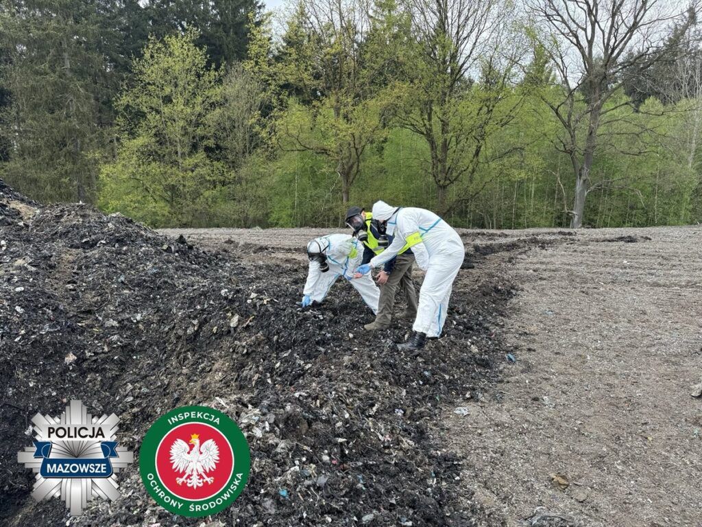 Dwaj mężczyźni w kombinezonach ochronnych przeszukują teren z odpadami w lesie.