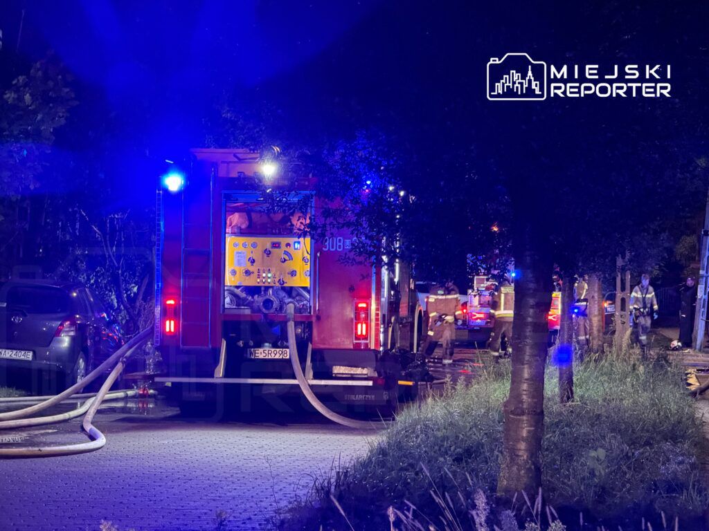 Straż pożarna z włączonymi sygnałami świetlnymi pracuje na ulicy, rozkładając węże gaśnicze.