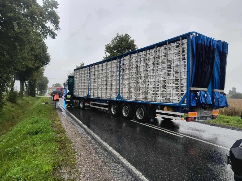 Kierowca ciężarówki rozładowuje towar na poboczu drogi w deszczową pogodę, obok pojazdu straży pożarnej.