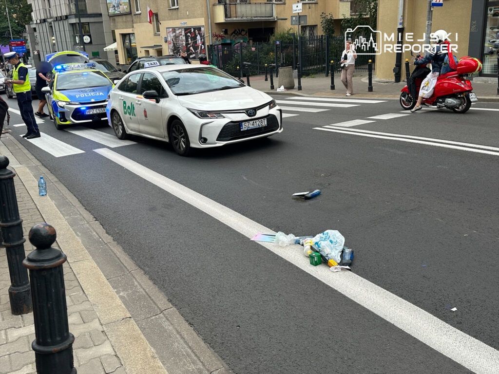 Na ulicy zatrzymane są dwa samochody, w tym taksówka i auto policyjne z włączonymi sygnalizatorami. Na jezdni leży porzucony zbiór odpadów.
