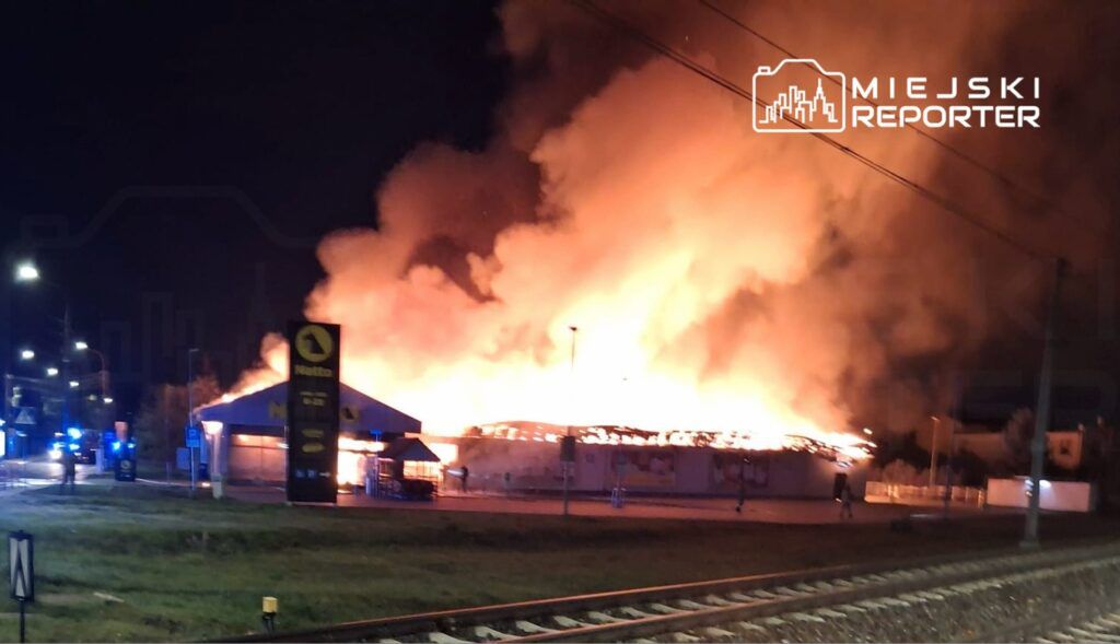 Intensywny pożar strawił budynek handlowy, otoczony przez dym i ogień, w nocy przy ulicy z samochodami strażackimi.