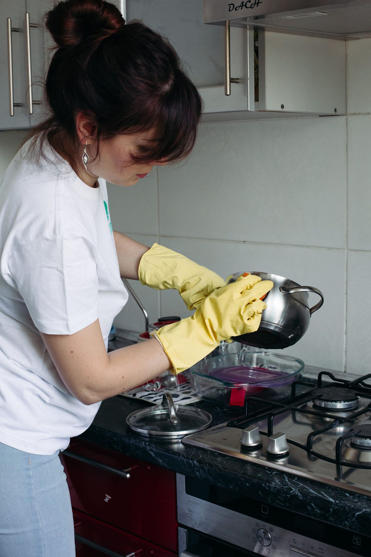 Close up of woman hands in white t shirt and protective yellow gloves washing dirty saucepan with sponge. Cooker, bowl and goblet on background. Concept of household chores.