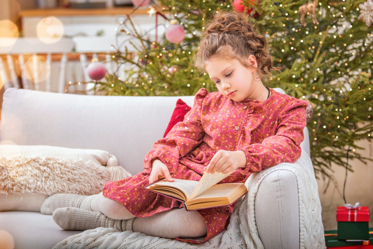 A cute little girl reads book at home in lived-in interior near a Christmas tree against the backdrop of burning fireplace.