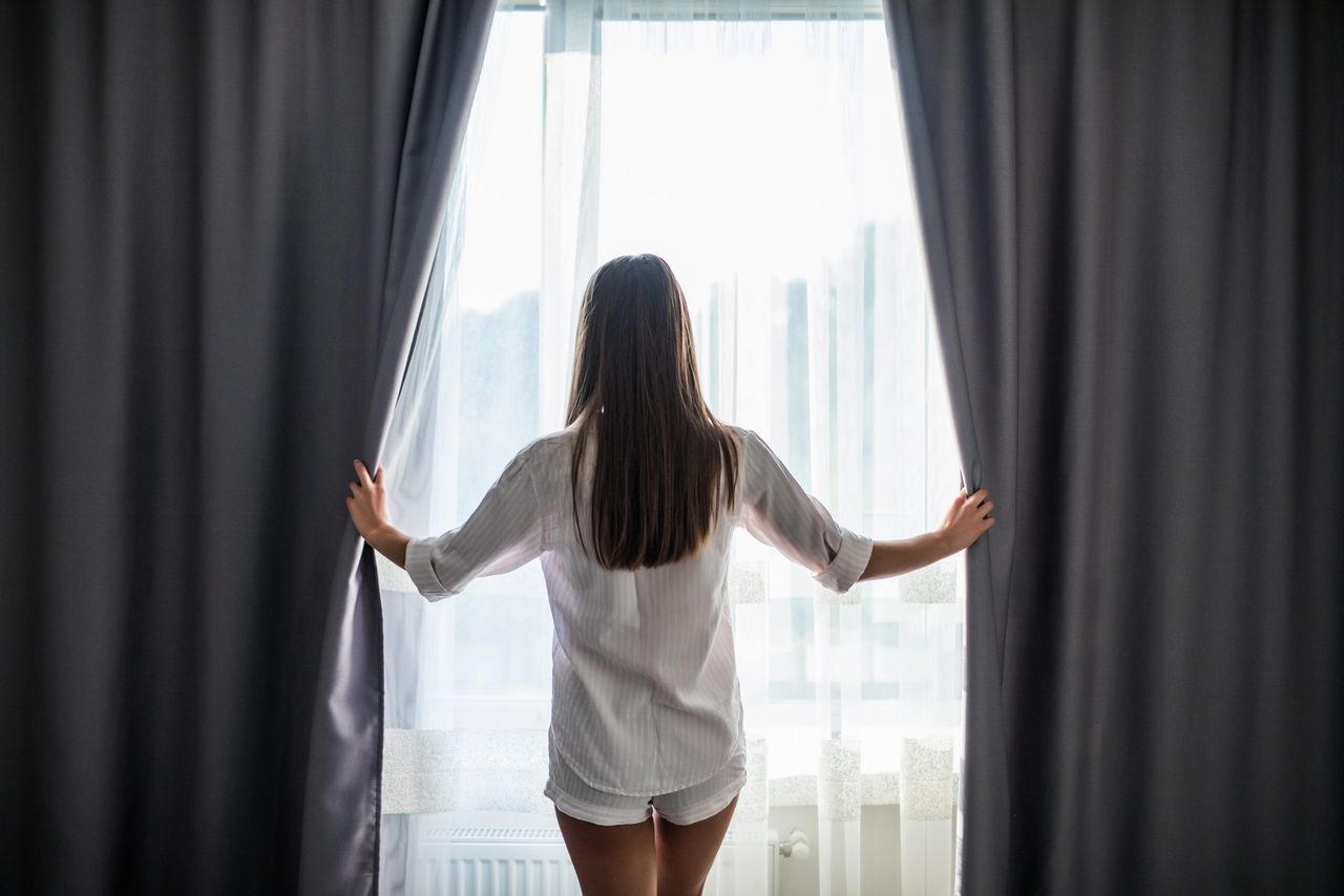 Beautiful woman opening curtains and looking through the window