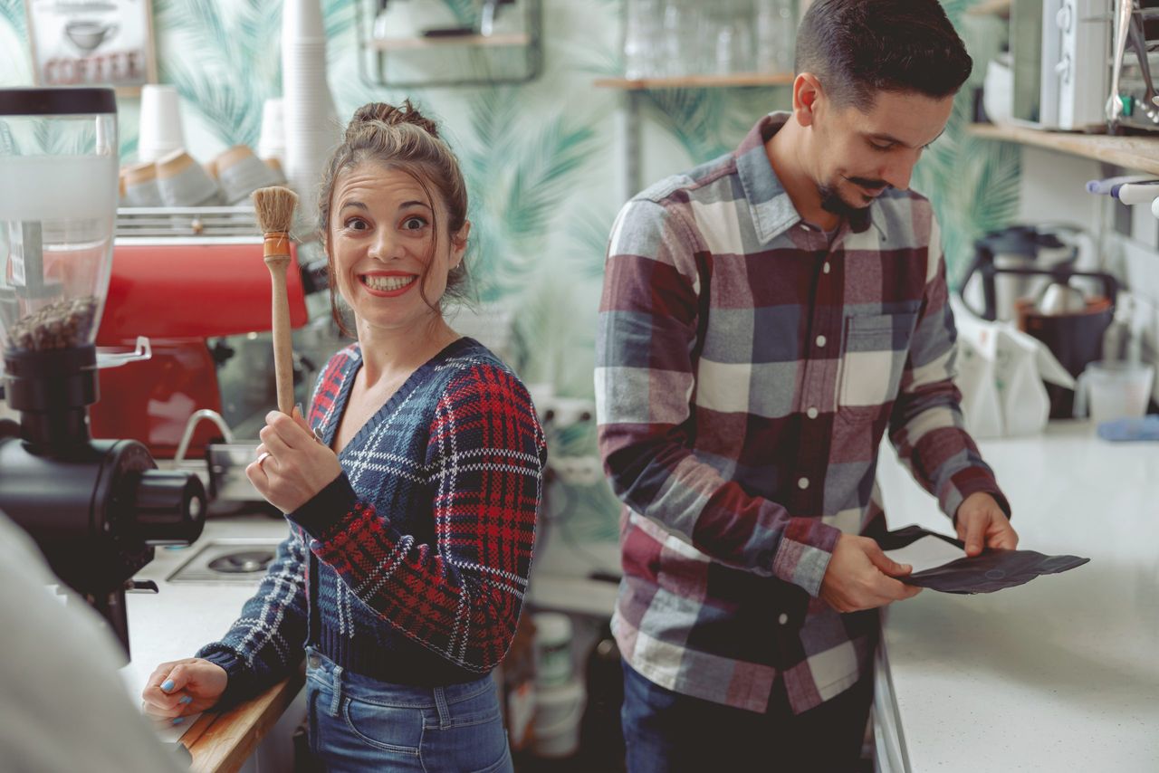 Smiling barista girl holding surface cleaning brush looking at camera and man standing nearby holding coffee package