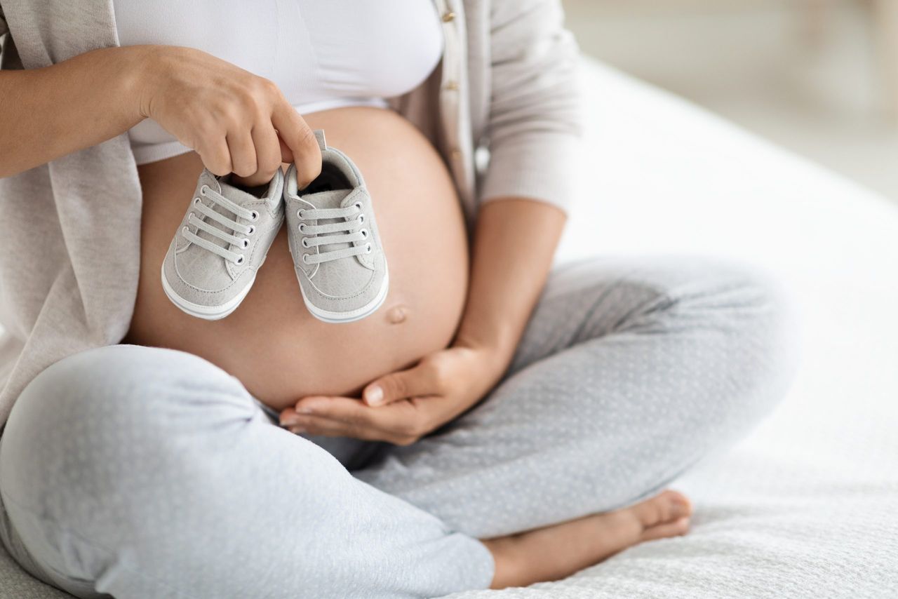Cropped of pregnant woman in comfy homewear sitting on bed at home, holding cute baby shoes on her big tummy, unrecognizable expecting mother waiting for baby child, copy space