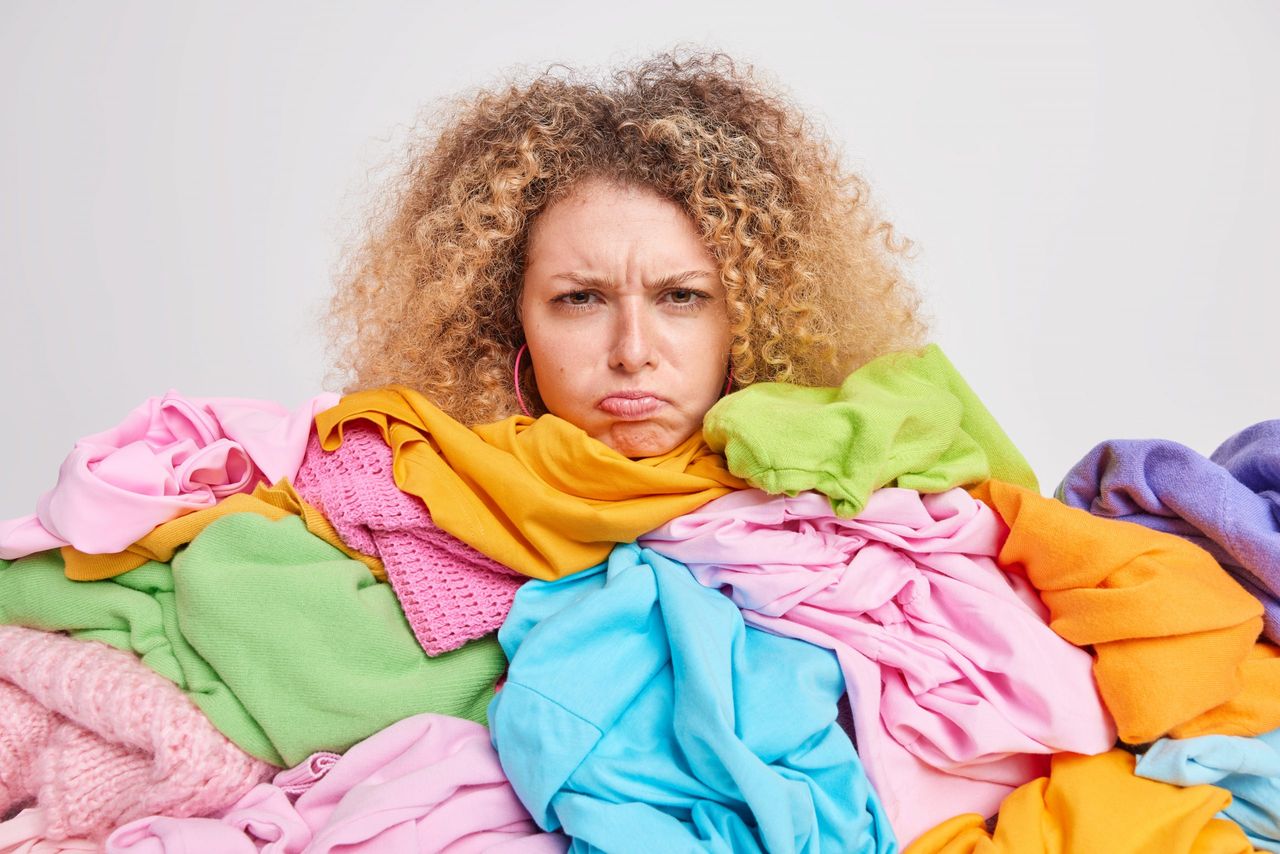 Discontent young European woman with curly hair has grumpy expression overloaded with colorful clothes upset to have much house work has to fold washed clothing isolated over white background