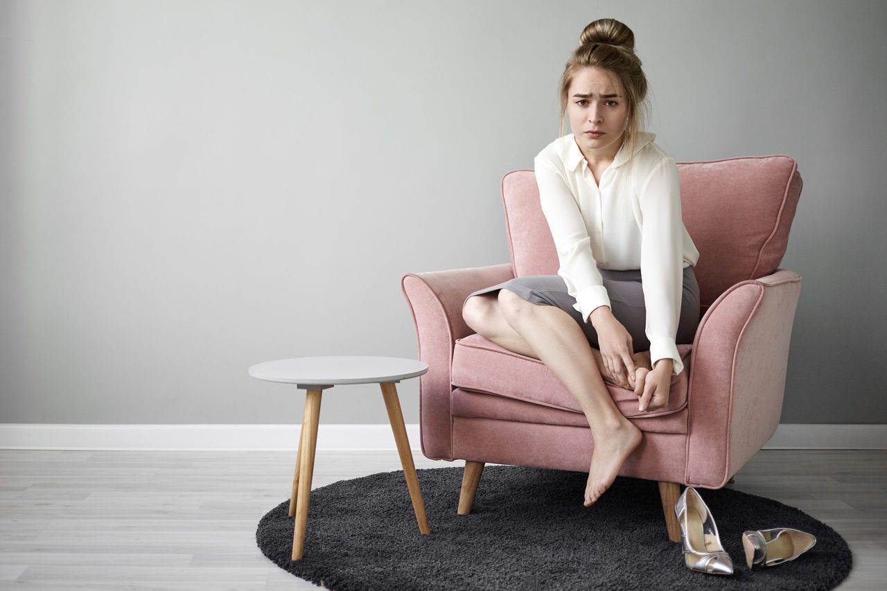 Modern hardworking young woman feeling tired after a long walk in high heeled shoes, sitting barefoot in an armchair, massaging her feet, with a painful and frustrated facial expression. Health and wellness.